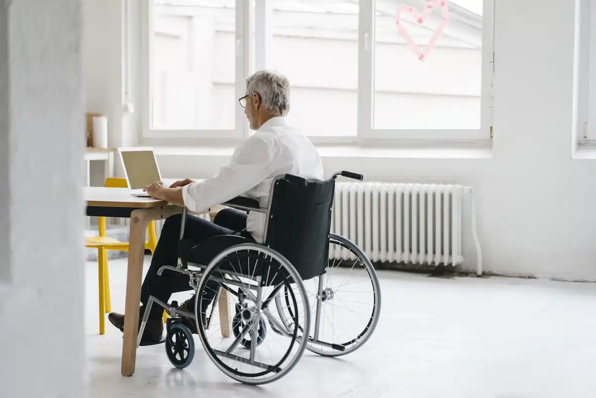 Cette image montre un individu dans un fauteuil roulant, entouré de dossiers médicaux et de documents administratifs. Il semble concentré et déterminé, peut-être en train de préparer une demande de reconnaissance de handicap auprès de la MDPH.
